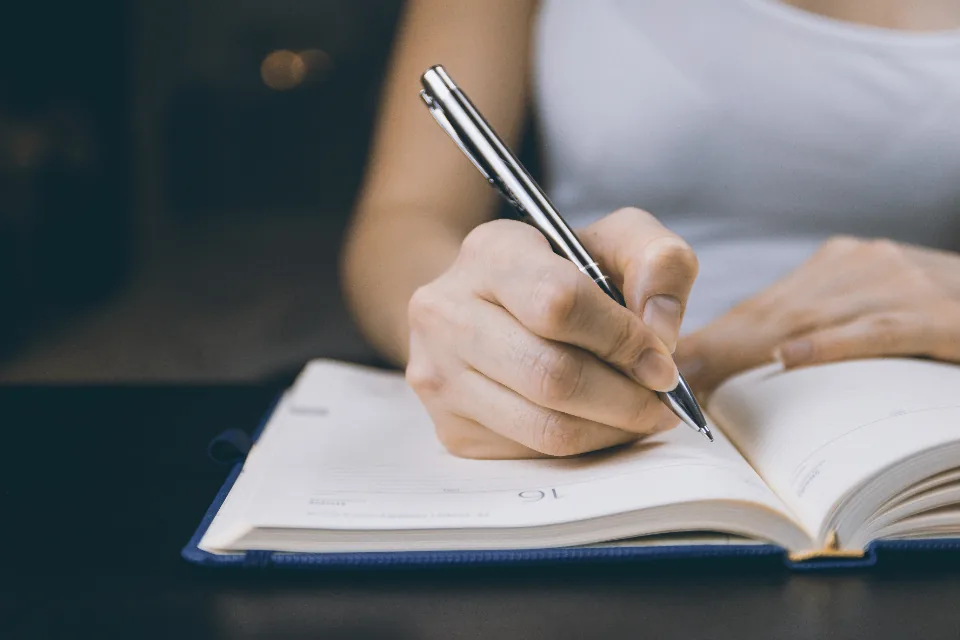 Une main écrit à la plume dans un carnet ouvert, sur un bureau sombre